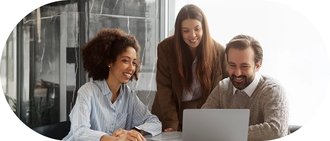 Grupo de três pessoas, duas mulheres e um homem, todos olhando para um notebook enquanto sorriem, vestindo roupas de negócios (suéteres, camisas sociais, etc.).