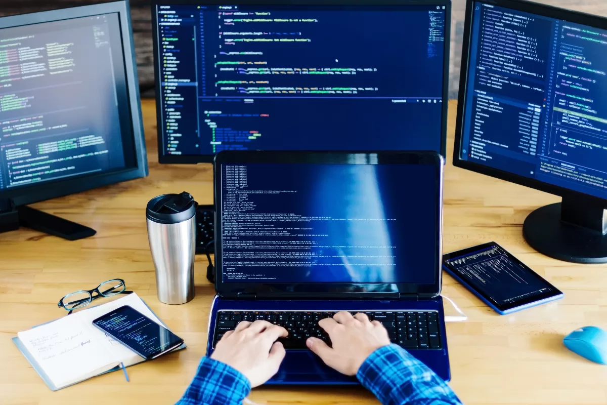 Hands typing in a laptop surrounded by computer screens.