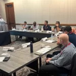 TestBooster's CEO standing and presenting at a conference room podium while several participants sit around U-shaped tables, listening and taking notes during a professional meeting, representing TestBooster at the Midsize Enterprise Summit