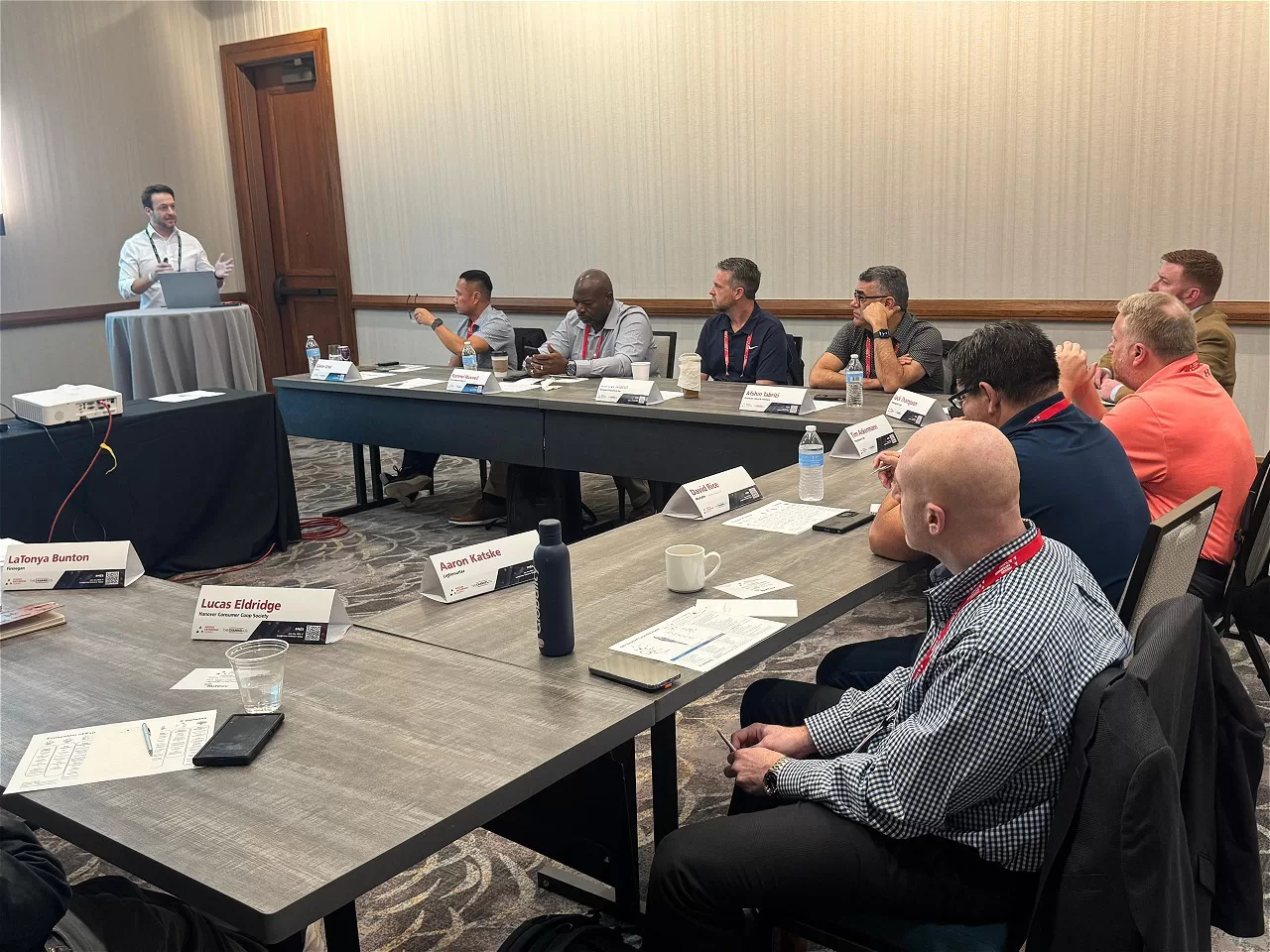 TestBooster's CEO standing and presenting at a conference room podium while several participants sit around U-shaped tables, listening and taking notes during a professional meeting, representing TestBooster at the Midsize Enterprise Summit
