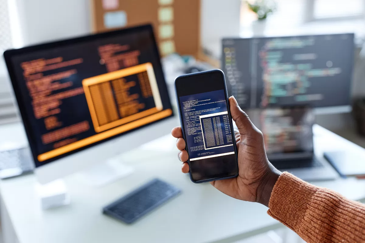 Person holding a smartphone displaying code, with multiple computer monitors in the background also showing programming interfaces