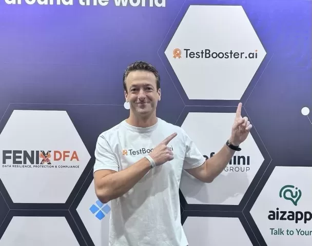 Juliano wearing a TestBooster.ai T-shirt standing in front of a conference backdrop with company logos, smiling and pointing with both hands at the TestBooster.ai logo.