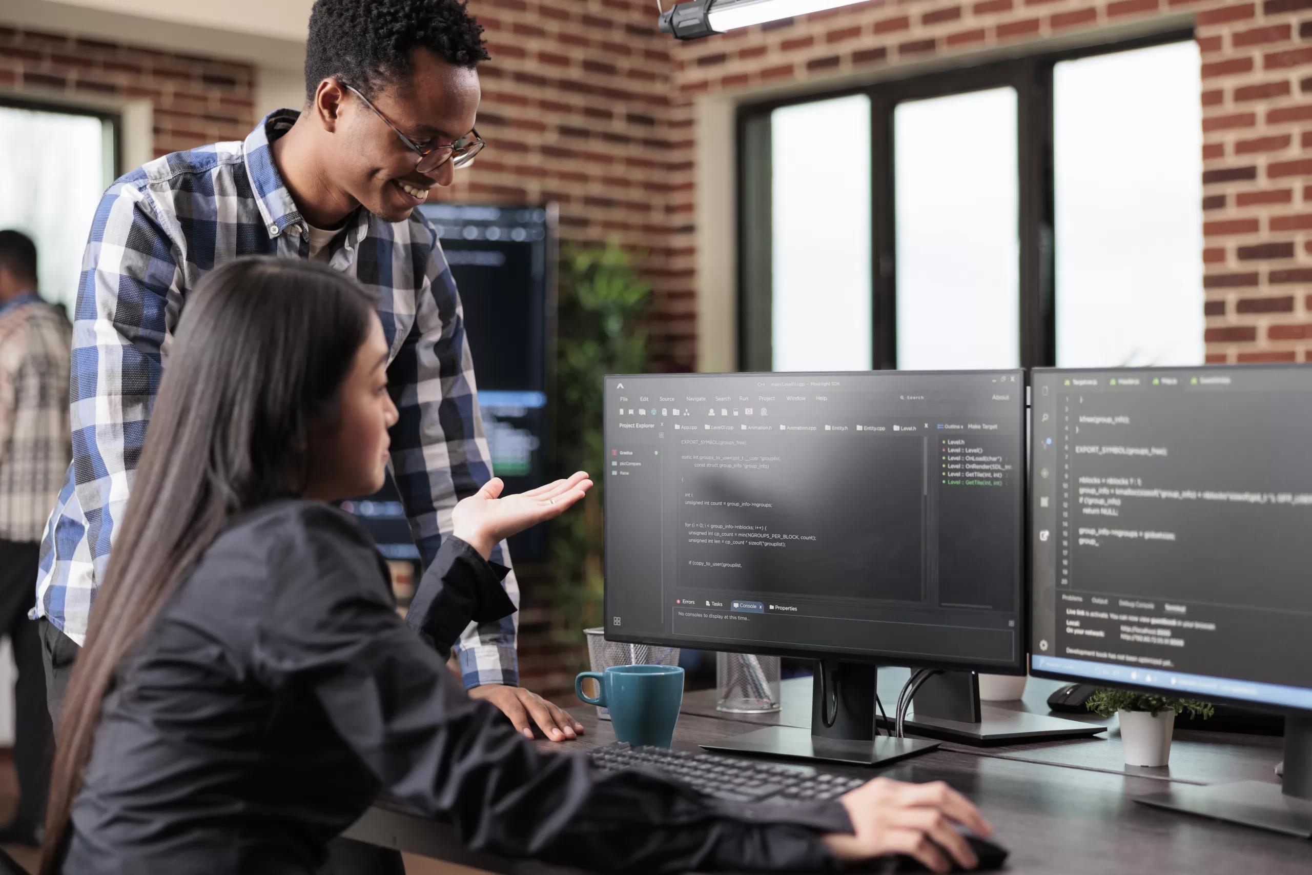 Two software developers discussing code on a dual-monitor setup in a modern office.