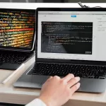 Person working on a laptop showing code, with an additional screen displaying colorful lines of programming code, representing