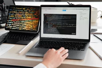 Person working on a laptop showing code, with an additional screen displaying colorful lines of programming code, representing