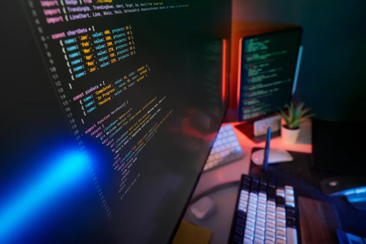 Um ambiente de trabalho com um monitor exibindo código em destaque, com luzes azuis e vermelhas. Um teclado mecânico e outro monitor secundário são visíveis no fundo.