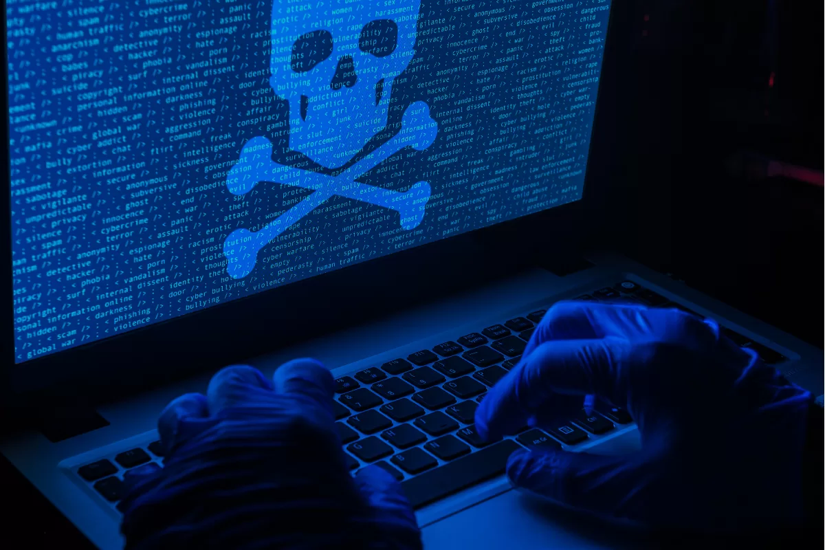Mãos com luvas digitando em laptop; caveira na tela simboliza ameaça de ataque cibernético.