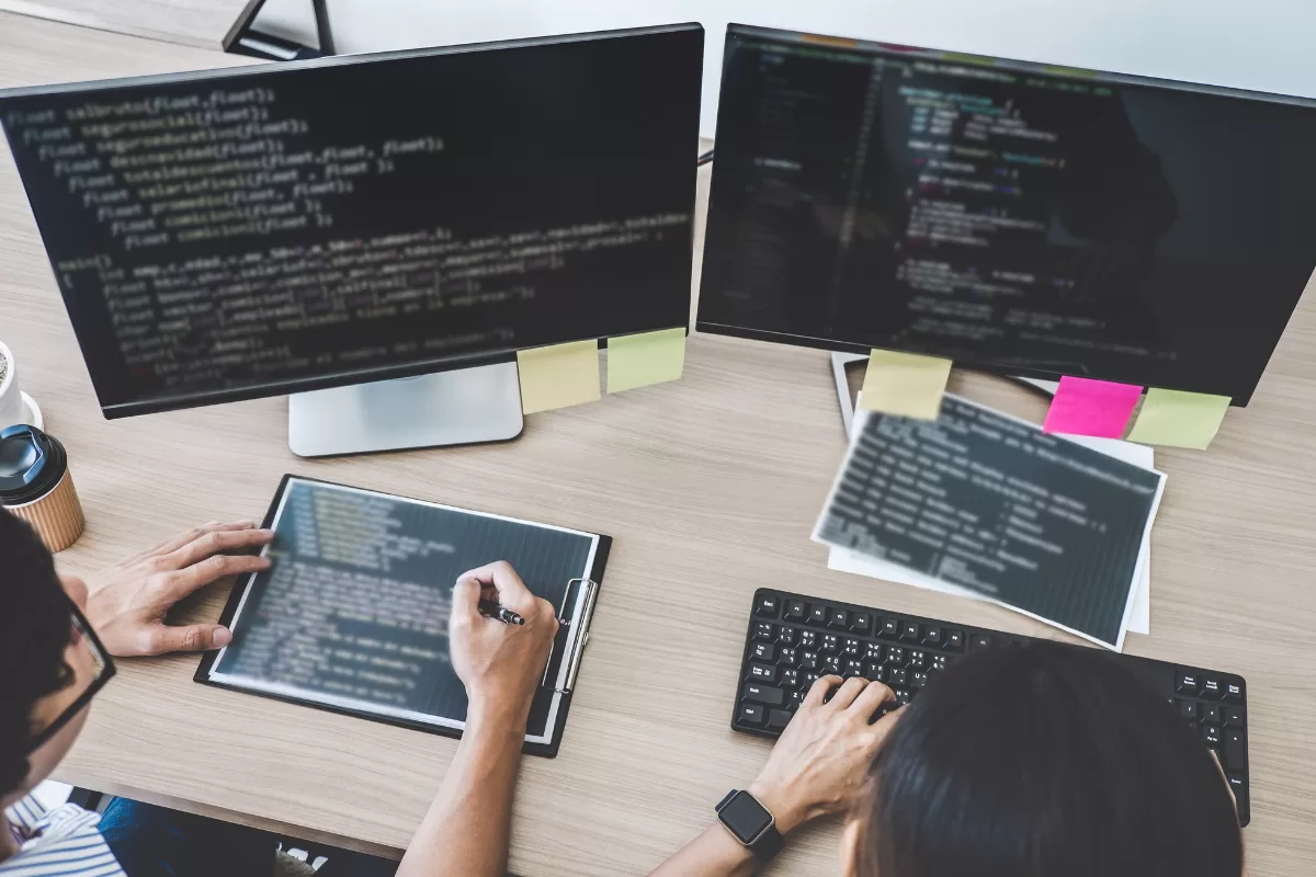 Two people working together at a desk, looking at two computer monitors. One person is writing on a clipboard while the other types on the keyboard, indicating software development collaboration.