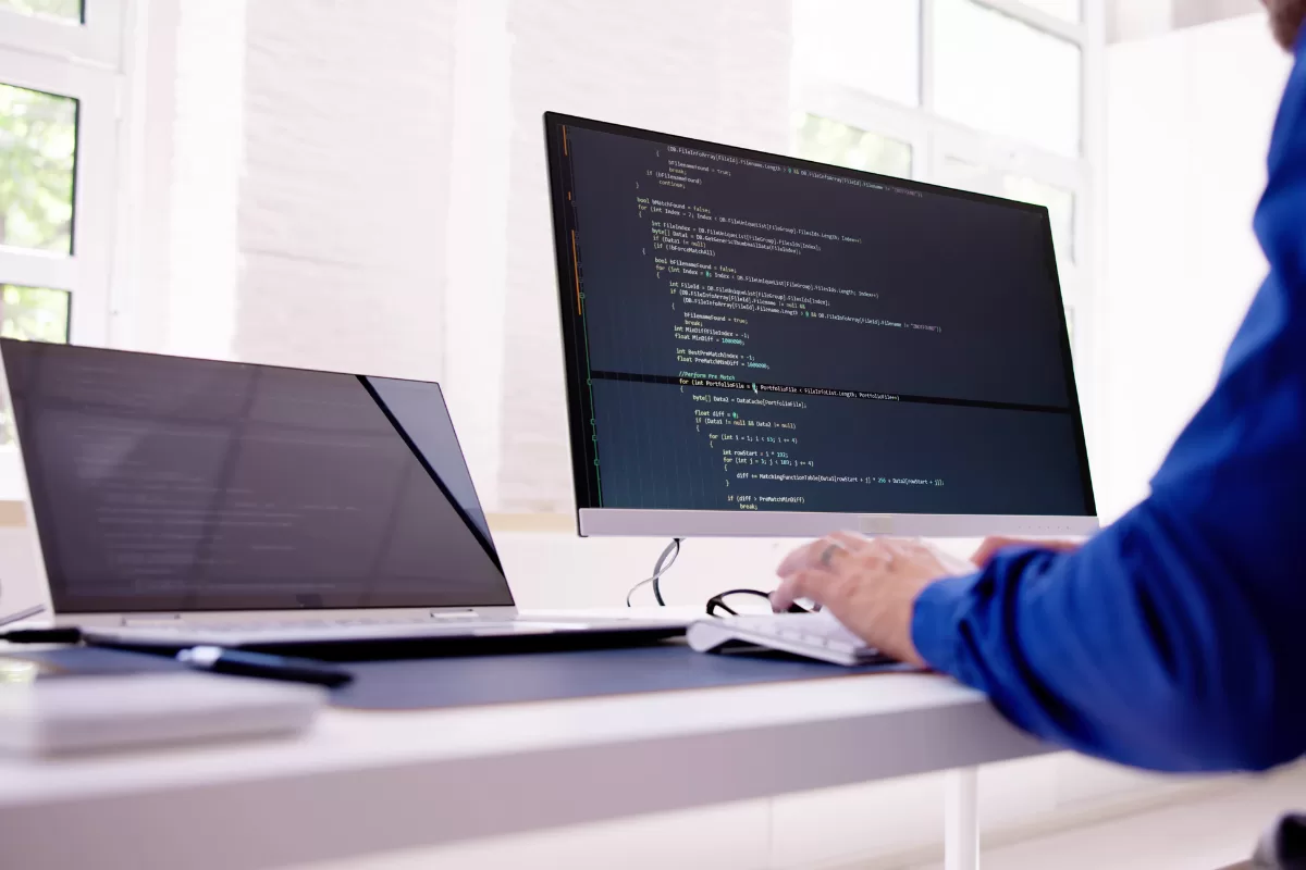 Desenvolvedor em camiseta azul trabalhando em monitor exibindo código de programação em ambiente de escritório iluminado