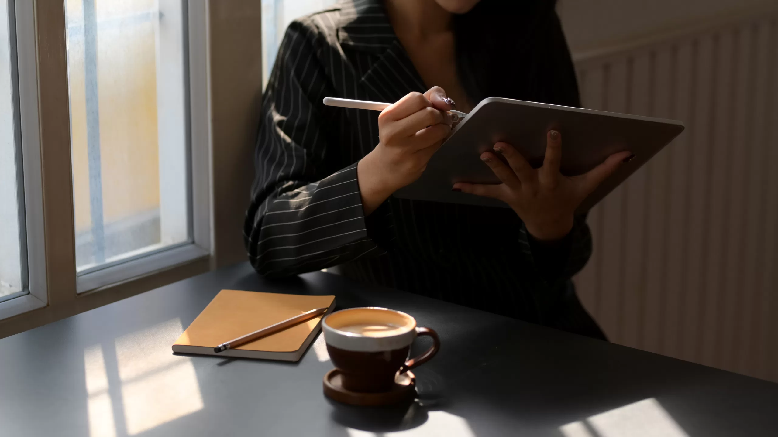 Close-up of a female freelancer in a suit jacket working on a digital tablet with a stylus, seated at a dark table with a cup of coffee nearby.
