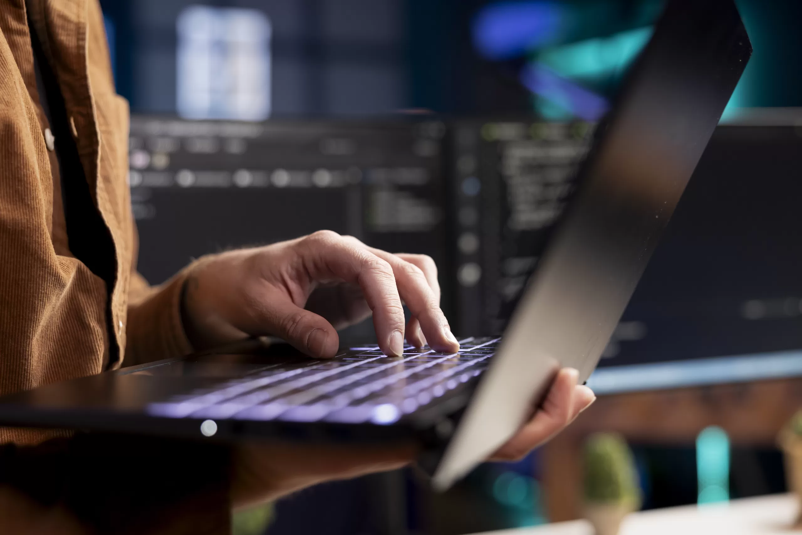 Person typing on laptop with multiple monitors in the background in a technical work environment