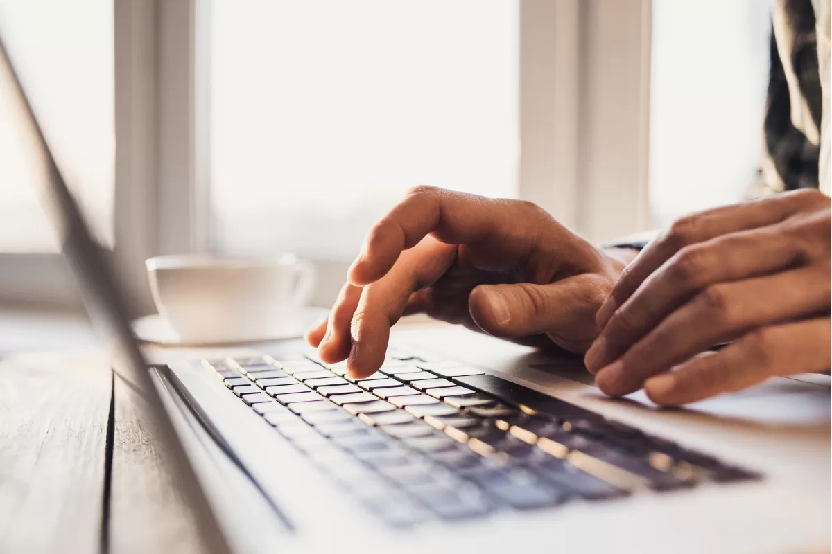 Hands typing on a laptop near a cup of coffee on a table.