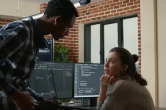 Dois profissionais de TI conversando em frente a monitores com códigos de programação em um escritório moderno.