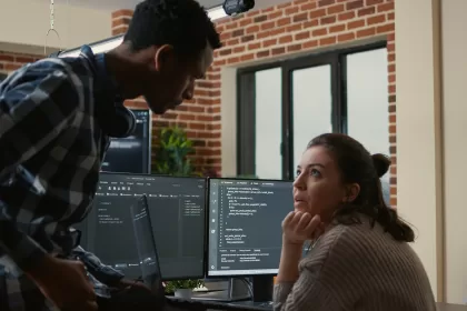 Dois profissionais de TI conversando em frente a monitores com códigos de programação em um escritório moderno.