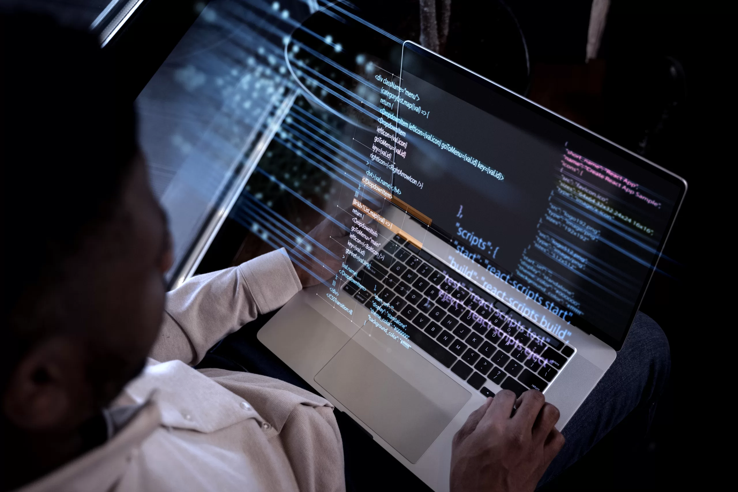 Top-down view of a person working on a silver laptop in the dark. A digital holographic interface with programming code (JSON and React) is overlaid on the screen and the user's hands, creating a futuristic tech effect.