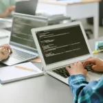 Desenvolvedora trabalhando em laptop com código na tela, realizando testes de software em ambiente colaborativo