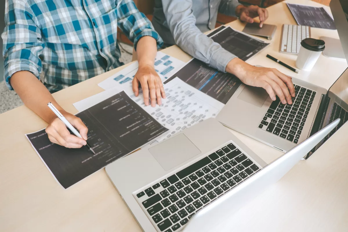 Equipe de desenvolvimento colaborando em testes de software com laptops e documentos de código, representando o trabalho em equipe na implementação de testes funcionais